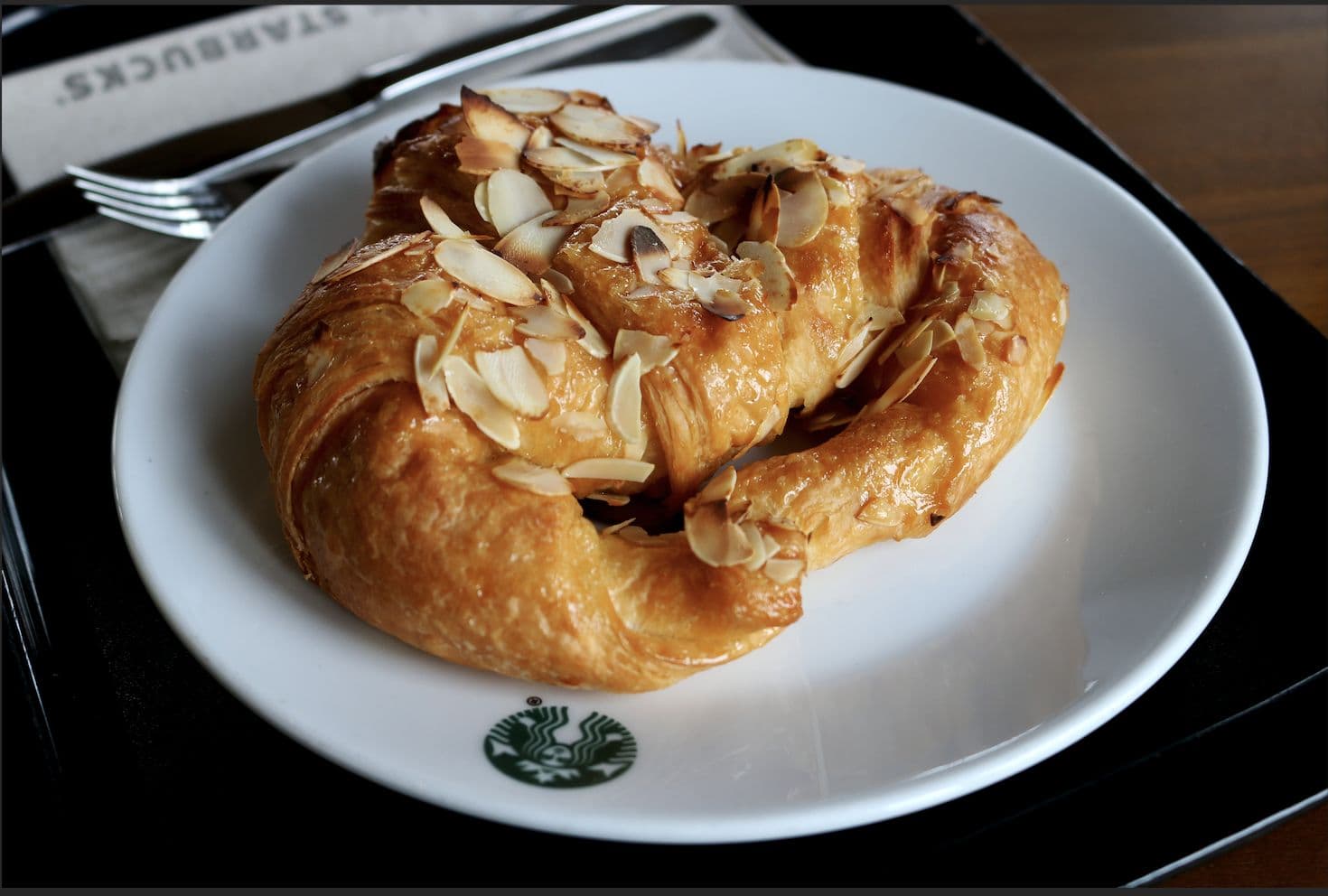 A glazed almond pastry on a white plate next to a fork on a dark table.