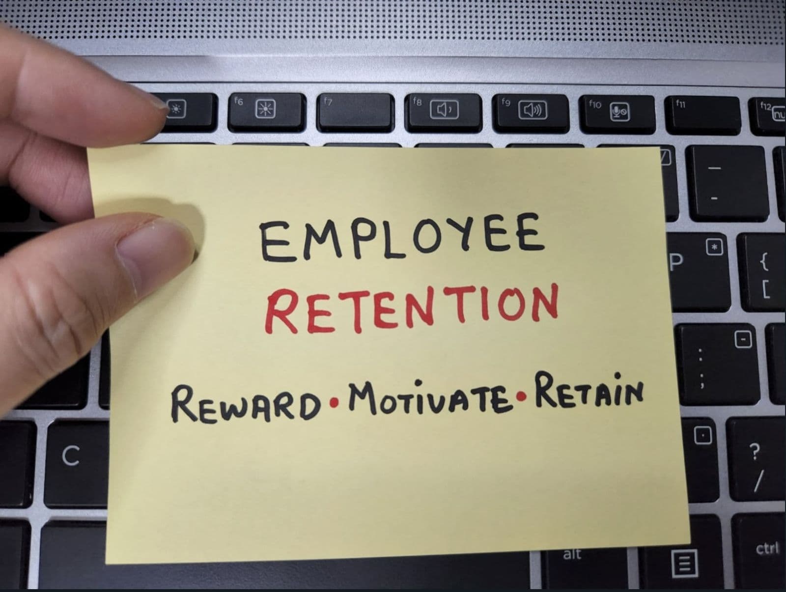 A hand holding a yellow sticky note that reads Employee Retention, Reward, Motivate, Retain, resting on a laptop keyboard.