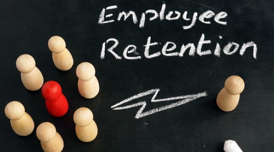 Wooden peg figures arranged on a blackboard next to the chalk-written words Employee Retention, with one figure in red.